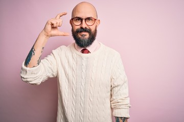 Handsome bald man with beard and tattoo wearing glasses and sweater over pink background smiling and confident gesturing with hand doing small size sign with fingers looking and the camera. Measure