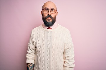 Handsome bald man with beard and tattoo wearing glasses and sweater over pink background making fish face with lips, crazy and comical gesture. Funny expression.