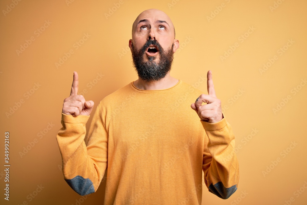 Poster handsome bald man with beard wearing casual sweater standing over yellow background amazed and surpr