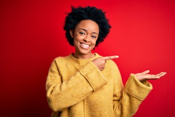 Young beautiful African American afro woman with curly hair wearing casual yellow sweater amazed and smiling to the camera while presenting with hand and pointing with finger.