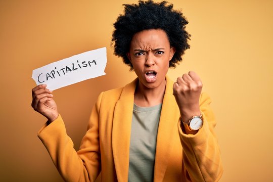 African American afro businesswoman with curly hair holding paper with capitalism message annoyed and frustrated shouting with anger, crazy and yelling with raised hand, anger concept