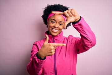 Young beautiful African American afro sportswoman with curly hair wearing pink sportswear smiling making frame with hands and fingers with happy face. Creativity and photography concept.