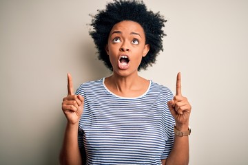 Young beautiful African American afro woman with curly hair wearing striped t-shirt amazed and surprised looking up and pointing with fingers and raised arms.
