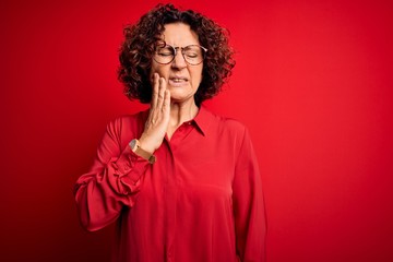 Fototapeta na wymiar Middle age beautiful curly hair woman wearing casual shirt and glasses over red background touching mouth with hand with painful expression because of toothache or dental illness on teeth. Dentist