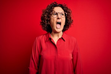 Fototapeta na wymiar Middle age beautiful curly hair woman wearing casual shirt and glasses over red background angry and mad screaming frustrated and furious, shouting with anger. Rage and aggressive concept.