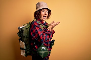 Plakat Middle age curly hair hiker woman hiking wearing backpack and water canteen using binoculars pointing aside with hands open palms showing copy space, presenting advertisement smiling excited happy