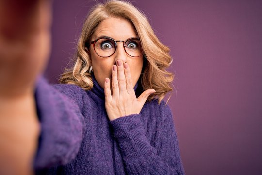 Middle age beautiful blonde woman wearing sweater and glasses make selfie by the camera cover mouth with hand shocked with shame for mistake, expression of fear, scared in silence, secret concept