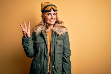 Middle age beautiful blonde skier woman wearing snow sportwear and ski goggles showing and pointing up with fingers number three while smiling confident and happy.
