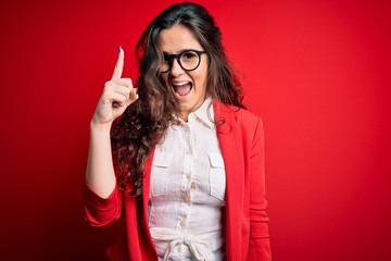 Young beautiful woman with curly hair wearing jacket and glasses over red background pointing finger up with successful idea. Exited and happy. Number one.