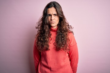 Young beautiful woman with curly hair wearing turtleneck sweater over pink background skeptic and...