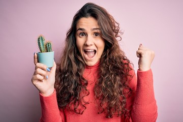 Young beautiful woman with curly hair holding small cactus over isolated pink background screaming proud and celebrating victory and success very excited, cheering emotion