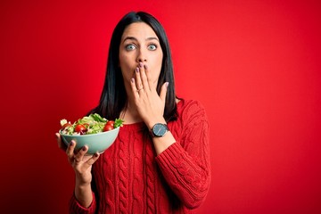 Young brunette woman with blue eyes eating healthy green salad over isolated red background cover mouth with hand shocked with shame for mistake, expression of fear, scared in silence, secret concept