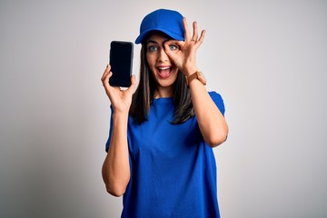 Young delivery woman with blue eyes wearing cap holding smartphone with happy face smiling doing ok sign with hand on eye looking through fingers