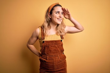 Young beautiful blonde woman wearing overalls and diadem standing over yellow background very happy and smiling looking far away with hand over head. Searching concept.