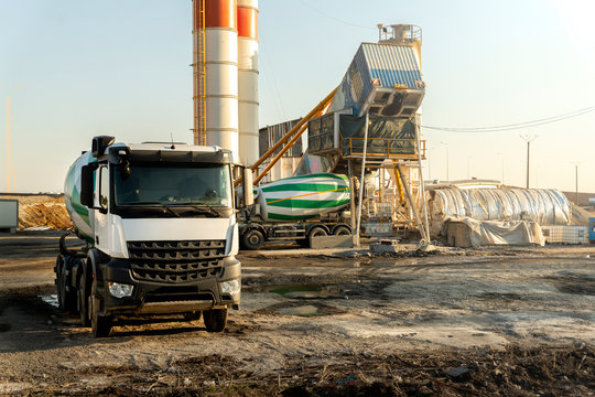 Row many modern big mixer trucks parked against mobile temporary concrete plant factory at new asphalt road construction site early morning day. Heavy machniery and industrial facilities background