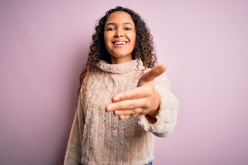 Young beautiful woman with curly hair wearing casual sweater standing over pink background smiling friendly offering handshake as greeting and welcoming. Successful business.