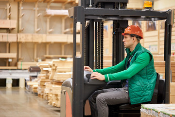 Fototapeta na wymiar confident warehouse worker sit inside of machine in cab. man in protective helmet and green uniform concentrated on work, wooden boards in the background