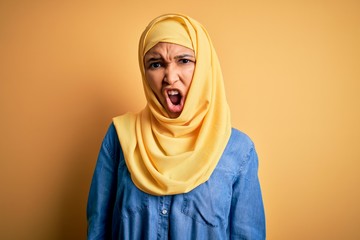 Young beautiful woman with curly hair wearing arab traditional hijab over yellow background In shock face, looking skeptical and sarcastic, surprised with open mouth