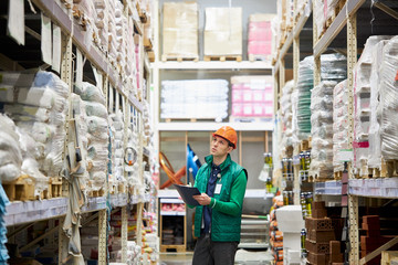 hard working caucasian male warehouse worker counts amount of goods in store and writes it in notebook, handsome guy in green unifrom and protective helmet concentrated on work
