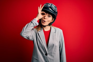 Young beautiful blonde motorcyclist woman wearing motorcycle helmet over red background doing ok gesture shocked with surprised face, eye looking through fingers. Unbelieving expression.