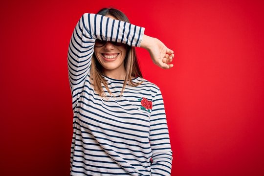 Young beautiful blonde woman with blue eyes wearing glasses standing over red background covering eyes with arm smiling cheerful and funny. Blind concept.