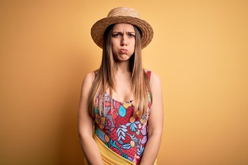 Young beautiful blonde woman wearing swimsuit and summer hat over yellow background puffing cheeks with funny face. Mouth inflated with air, crazy expression.