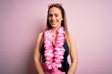 Young beautiful blonde woman wearing swimsuit and floral Hawaiian lei over pink background winking...