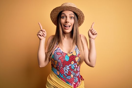 Young beautiful blonde woman wearing swimsuit and summer hat over yellow background smiling amazed and surprised and pointing up with fingers and raised arms.