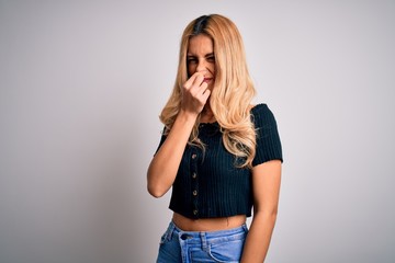 Young beautiful blonde woman wearing casual t-shirt standing over isolated white background smelling something stinky and disgusting, intolerable smell, holding breath with fingers on nose. Bad smell