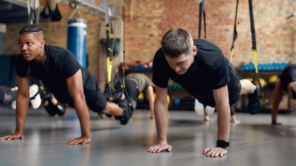 Motivated People Only. Caucasian man stretching his body while doing fitness TRX training exercises at industrial gym. Straps, group workout concept - obrazy, fototapety, plakaty