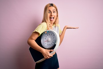 Young beautiful blonde sporty woman on diet holding weight machine over pink background very happy and excited, winner expression celebrating victory screaming with big smile and raised hands