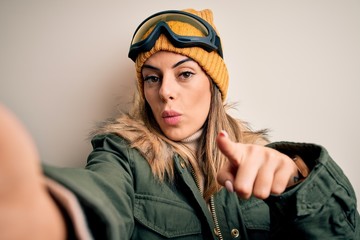Young brunette skier woman wearing snow clothes and ski goggles make selfie by camera pointing with finger to the camera and to you, hand sign, positive and confident gesture from the front