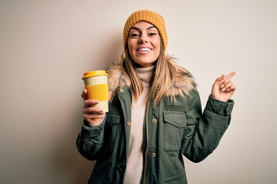 Young beautiful brunette woman wearing snow clothes drinking cup of coffee very happy pointing with hand and finger to the side