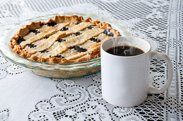 White coffee mug and blueberry pie still life on a lace tablecloth.  Styled product photo with copy space on the coffee cup for custom text or graphics.  Steaming hot black coffee and pie dessert.