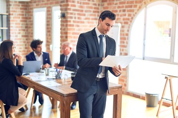 Group of business workers smiling happy and confident working together in a meeting. One of them, standing with smile on face reading documents at the office.