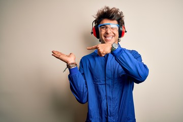 Young constructor man wearing uniform and earmuffs over isolated white background amazed and smiling to the camera while presenting with hand and pointing with finger.