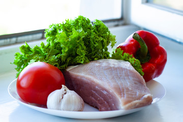 A piece of raw pork served on a white plate with sweet pepper, lettuce, tomato and garlic. Healthy eating and cooking at home concept. Cooking of meat dish, tasty dinner, lunch