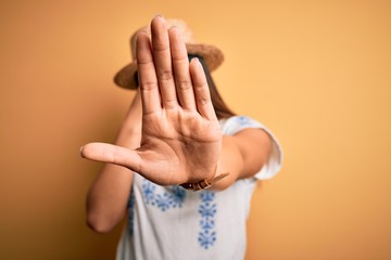 Young beautiful asian girl wearing casual t-shirt and hat standing over yellow background covering eyes with hands and doing stop gesture with sad and fear expression. Embarrassed and negative