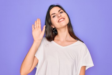 Fototapeta premium Young beautiful brunette woman wearing casual white t-shirt over purple background Waiving saying hello happy and smiling, friendly welcome gesture