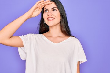 Young beautiful brunette woman wearing casual white t-shirt over purple background very happy and smiling looking far away with hand over head. Searching concept.