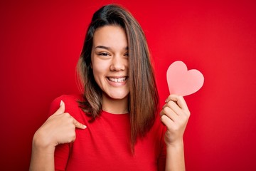 Young beautiful brunette romantic girl holding red paper heart shape over isolated background with surprise face pointing finger to himself