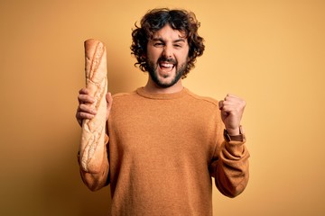 Young handsome man with curly hair and beard holding homemade healthy bread screaming proud and...