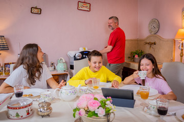 Happy family is having breakfast together at home in the kitchen - Mother, son, father and daughter having typical italian breakfast - Children are watching the tablet and the father is making coffee