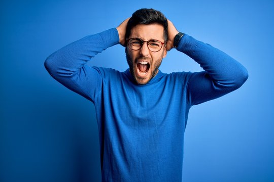 Young handsome man with beard wearing casual sweater and glasses over blue background Crazy and scared with hands on head, afraid and surprised of shock with open mouth