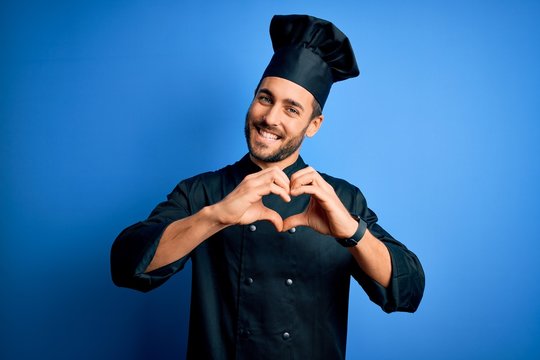 Young handsome chef man with beard wearing cooker uniform and hat over blue background smiling in love doing heart symbol shape with hands. Romantic concept.