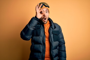 Young handsome skier man with beard wearing snow sportswear and ski goggles doing ok gesture shocked with surprised face, eye looking through fingers. Unbelieving expression.