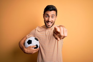 Handsome player man with beard playing soccer holding footballl ball over yellow background pointing to you and the camera with fingers, smiling positive and cheerful