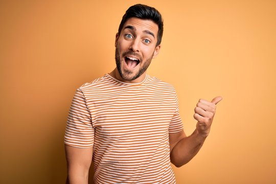 Young handsome man with beard wearing casual striped t-shirt over yellow background smiling with happy face looking and pointing to the side with thumb up.