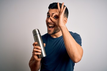 Young handsome tourist man drinking thermo with water over isolated white background with happy face smiling doing ok sign with hand on eye looking through fingers