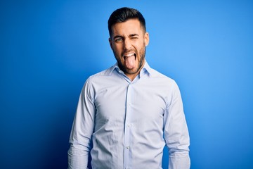 Young handsome man wearing elegant shirt standing over isolated blue background sticking tongue out happy with funny expression. Emotion concept.